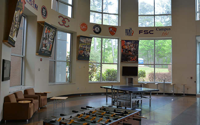 image of the gaming zone second area, white wall with several windows logos for CSIR, NSLS, CEC, SAC, AmeriCorps, SGA, The Bent Tree, FSC, & Campus Life as well as a ping pong table, a foosball table, two chairs, and a large televeision