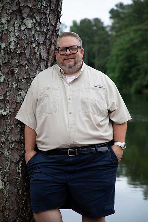 josh meddaugh leaning on a tree next to a lake