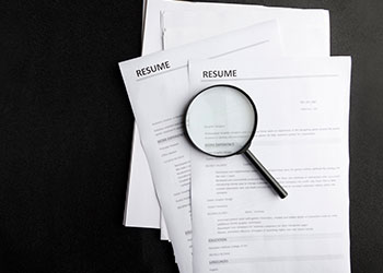 Magnifying glass over printed resumes on a dark background.