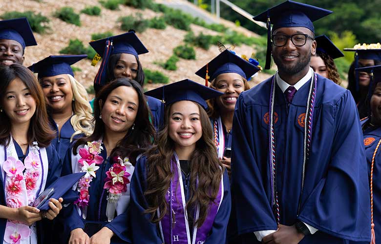 Group of graduates wearing blue caps and gowns pose together outdoors before commencement. Several wear honor cords, medals, and leis, highlighting academic achievements. The graduates stand closely for a commemorative photo celebrating graduation day.