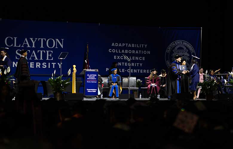 Graduation ceremony in progress on a Clayton State University commencement stage. Graduates and university officials participate as a diploma is presented. Blue university banners, ceremonial seating, flags, and stage décor are visible before a large audience.
