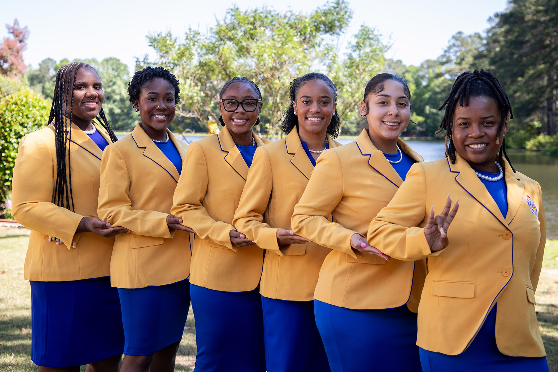 Members of Rho Lambda Chapter of Sigma Gamma Rho standing in a line, holding ther person in front of each members' raised elbow, while wearing gold jackets, blue dresses, and a string a of pearls